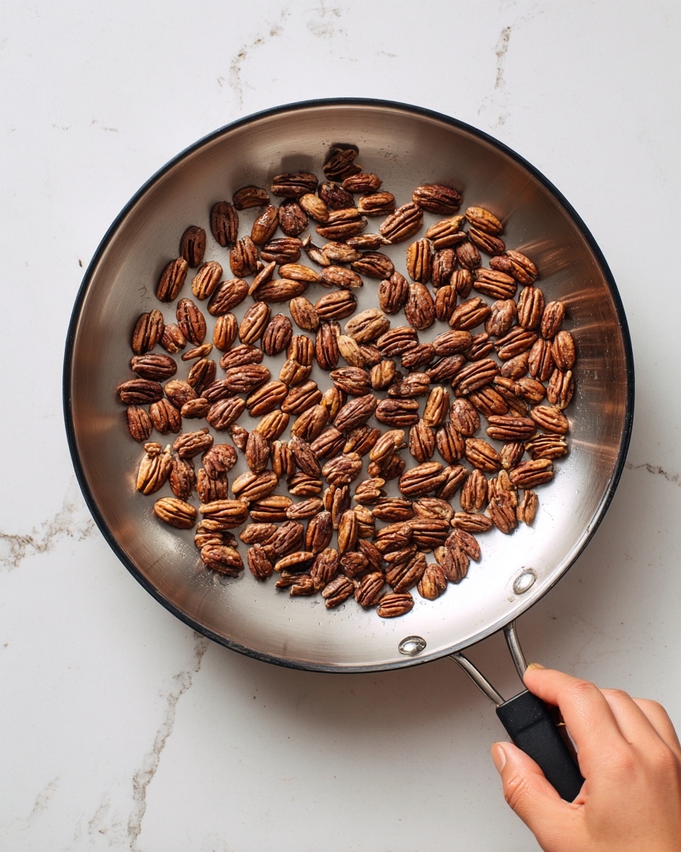A silver frying pan with a black inside is placed on a white marbled surface. Inside the pan, there are many medium brown pecan halves spread in a single layer. A woman's hand is holding the pan handle on the bottom right side. The overall look is simple, with the nuts' textured shells contrasting with the smooth pan surface. photo taken with an iphone --ar 4:5 --v 7