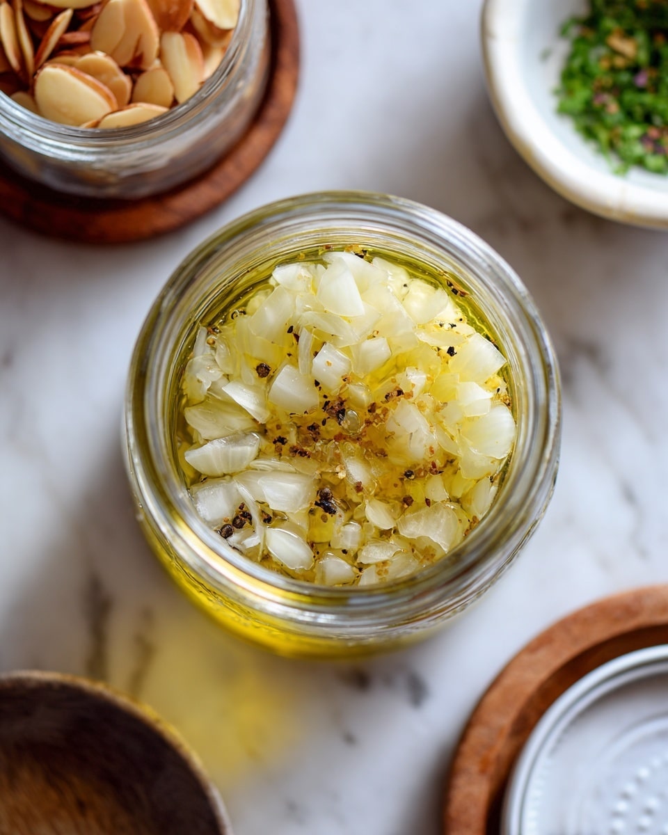 A clear glass jar viewed from above with three visible layers inside: a bottom layer of finely chopped light yellow shallots, a middle layer of bright golden oil, and a top layer showing a few pieces of thinly sliced shallots and black pepper specks. Around the jar, there is a white bowl with green chopped herbs at the top right, a glass container filled with toasted, light brown almond slices on the left placed on a wooden coaster, and the white jar lid at the bottom right. All items are set on a white marbled surface. photo taken with an iphone --ar 4:5 --v 7