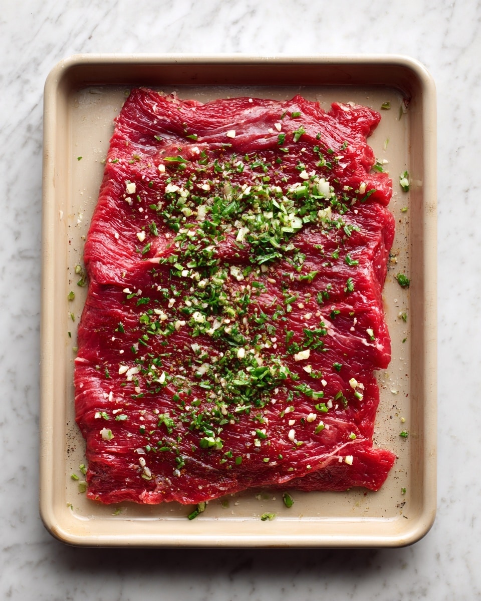 A large flat piece of raw red meat spread evenly on a beige baking tray. The meat is bright red with some darker and lighter areas of texture. On top, there is a scattered layer of finely chopped green herbs mixed with small bits of white garlic, covering most of the meat surface but not fully. The baking tray sits on a white marbled surface. photo taken with an iphone --ar 4:5 --v 7