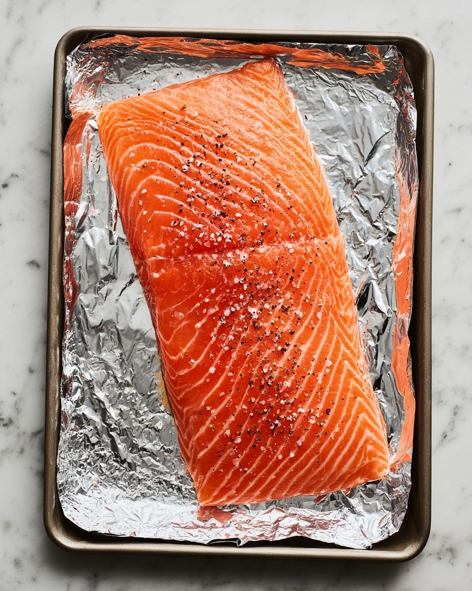 A large piece of raw salmon with bright orange color and white streaks lays flat on a sheet of shiny silver foil that covers the surface of a metal baking tray. The salmon is sprinkled with coarse salt and ground black pepper, showing small flecks over its smooth and slightly glossy texture. The tray edges frame the foil and fish against a white marbled background. photo taken with an iphone --ar 4:5 --v 7