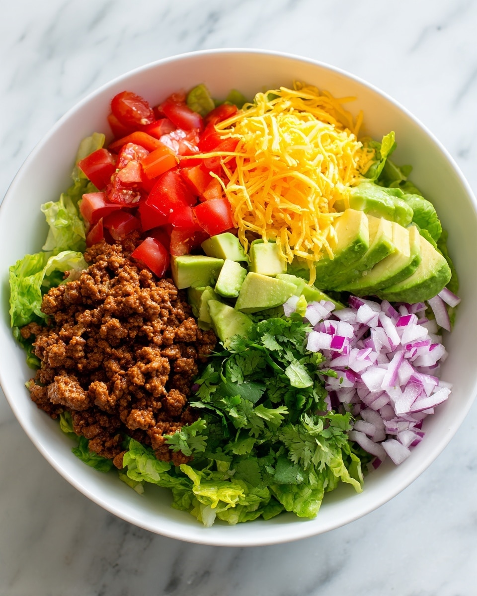 A large white bowl with speckles holds a colorful mixed salad. The salad has many layers including bright green chopped lettuce, shredded orange cheese, small red tomato pieces, and bits of brown cooked ground meat. A creamy dressing with green herbs is visible, coating some parts of the salad. A spoon is lifting a big portion of the salad, showing the fresh and textured layers clearly. The bowl sits on a white marbled surface, with a blurry background showing a white plate with light-colored chips. Photo taken with an iphone --ar 4:5 --v 7