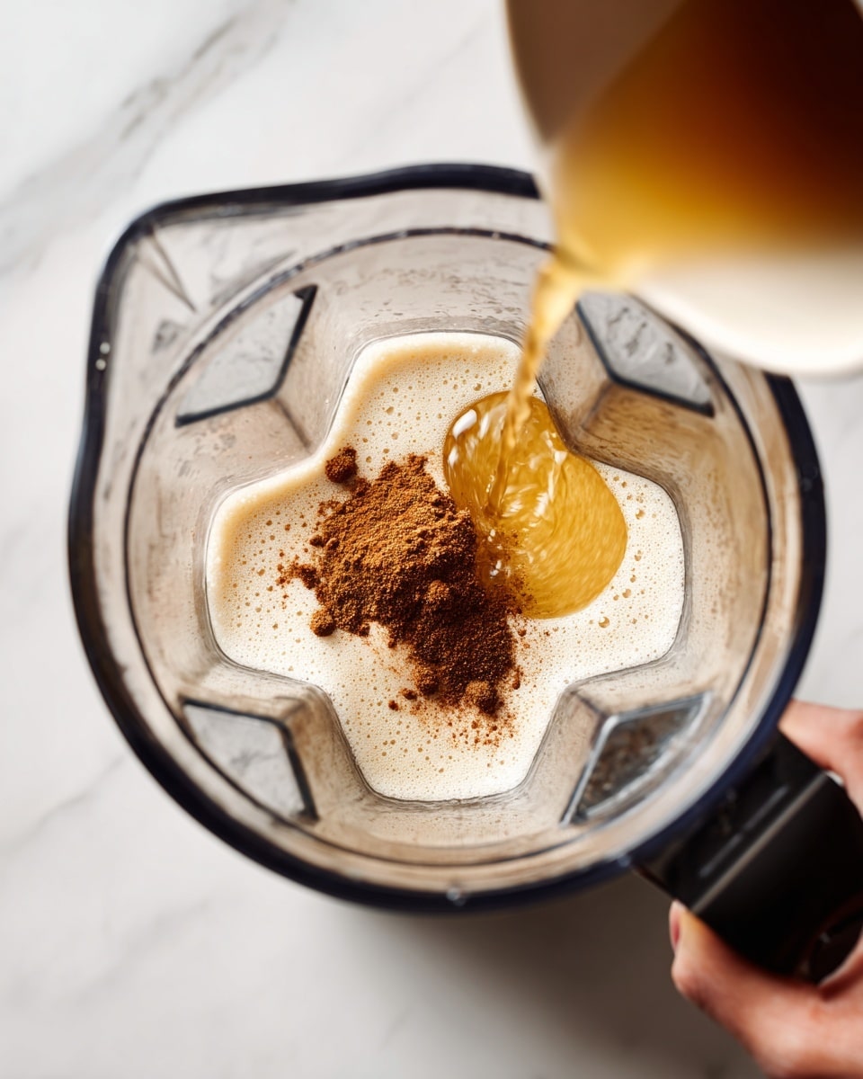 A clear blender jar is shown from above on a white marbled surface. Inside the blender, there is a frothy light cream layer at the bottom, topped with a small mound of finely ground brown spice in the center. A woman's hand on the right side pours a thin stream of golden liquid into the blender, creating a slight ripple effect on the foam and spice layers. The blender itself is transparent with dark edges and small textured details inside. photo taken with an iphone --ar 4:5 --v 7