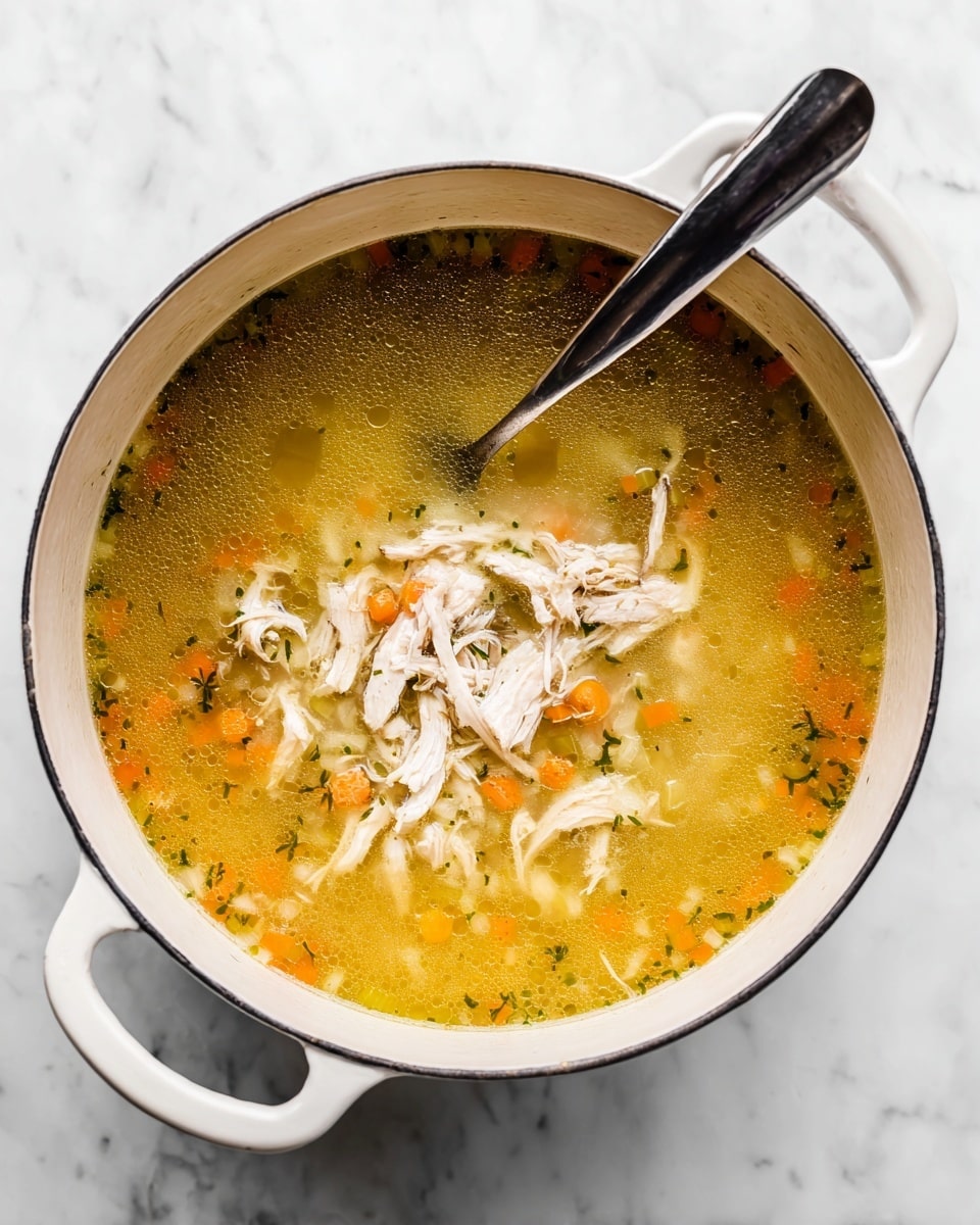 A white pot filled with clear golden broth that contains small diced vegetables visible through the liquid layer, topped with a layer of shredded white chicken pieces in the center. A black and silver spoon rests inside the pot, partially submerged in the broth on the right side. The pot has two handles and is set against a white marbled surface. photo taken with an iphone --ar 4:5 --v 7