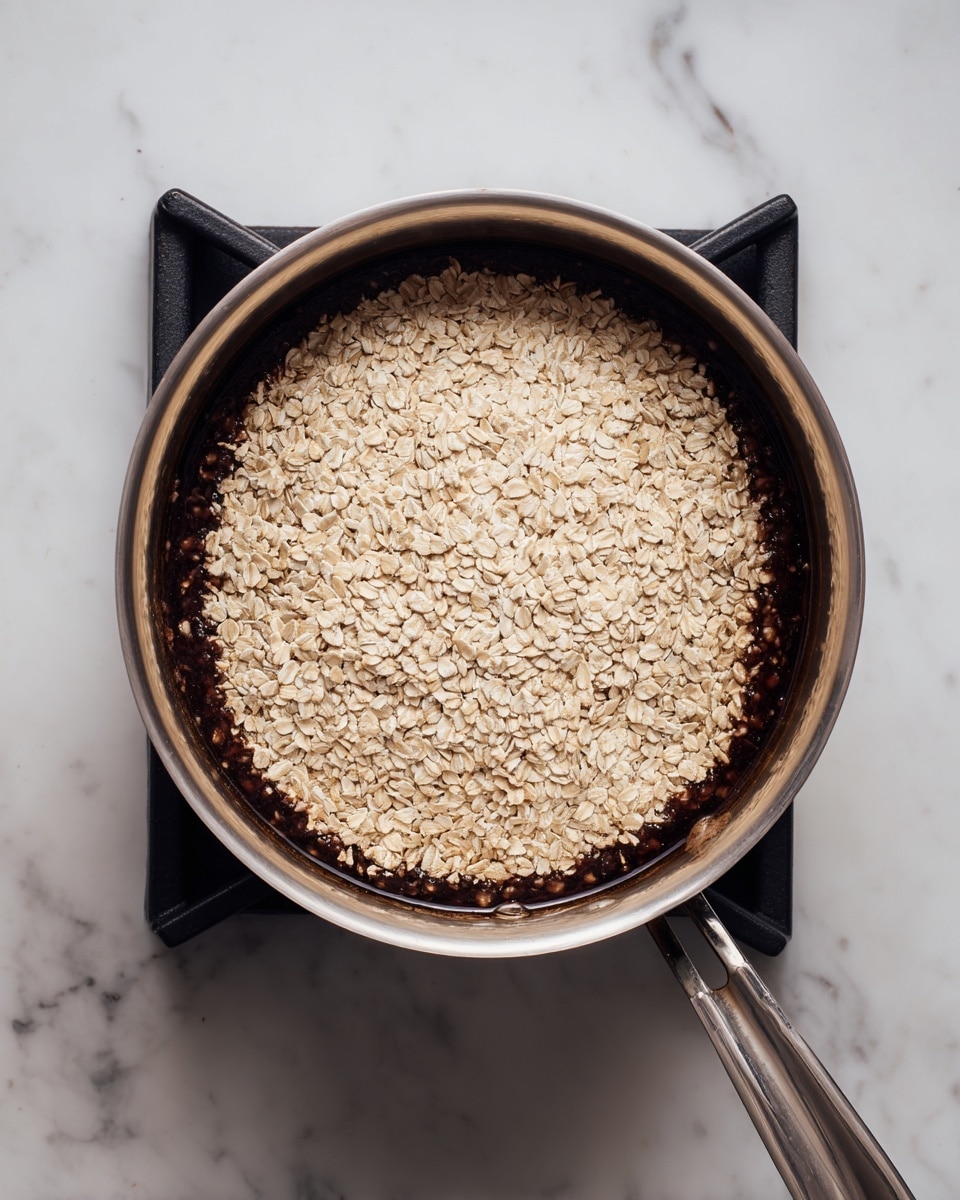 The image shows a metal saucepan on a stove with black grates, containing two visible layers: the bottom layer is dark brown and looks thick, filling the entire pan base, while the top layer is a large pile of light beige oats, spread evenly in a slightly rounded mound. The saucepan handle extends to the right, and the background is a white marbled texture. Photo taken with an iphone --ar 4:5 --v 7