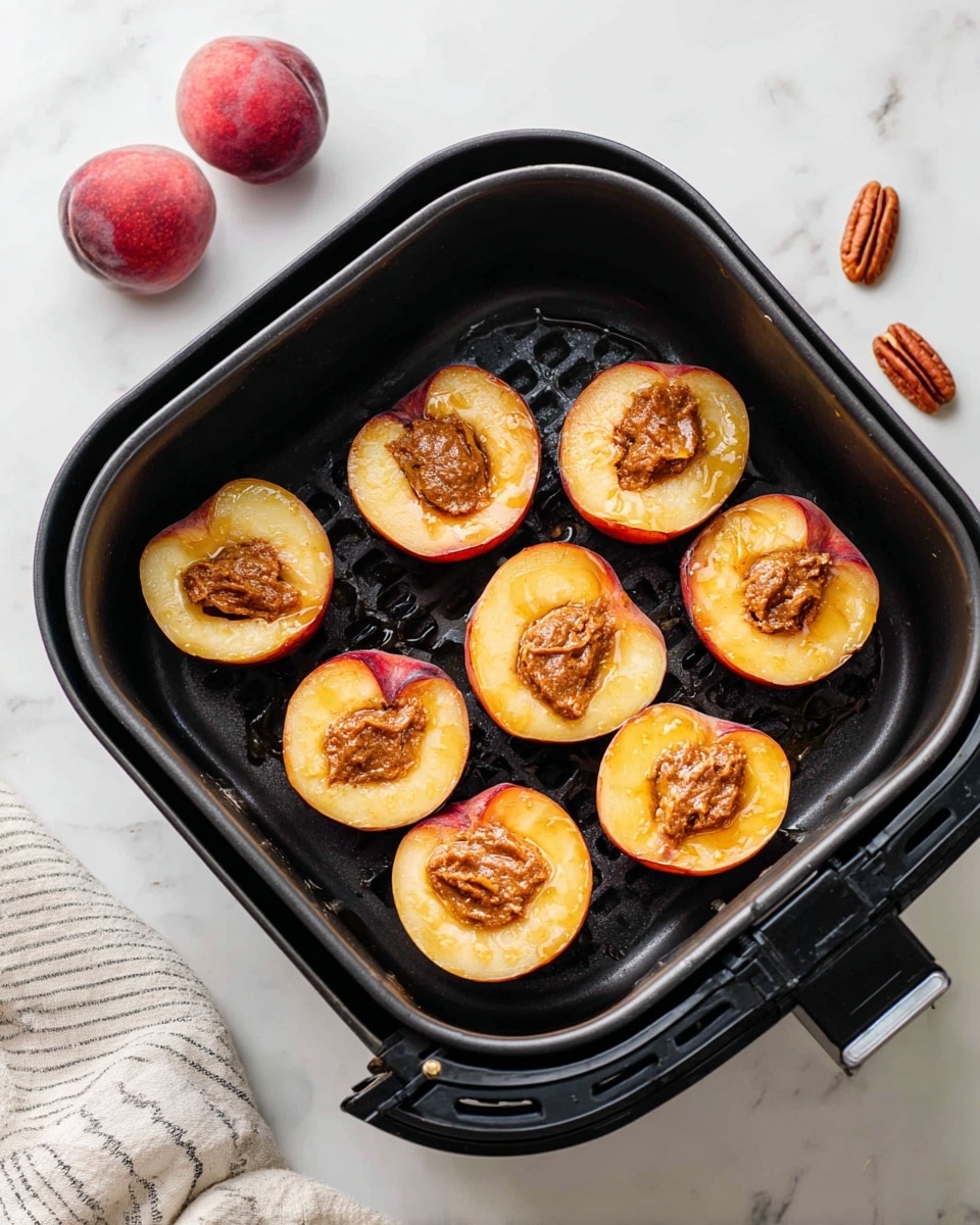 The image shows eight peach halves placed cut side up in a black air fryer basket on a white marbled surface. Each peach half has a dollop of brown almond butter filling in the center, with a drizzle of syrup or honey over it, adding a shiny texture. The peaches have a reddish-orange skin with a pale yellow inside, creating a contrast with the topping. Two reddish-brown whole pecans are visible on the white marbled surface near the air fryer. A white cloth with gray stripes is partially seen at the bottom left corner, adding a soft element to the image. Photo taken with an iphone --ar 4:5 --v 7