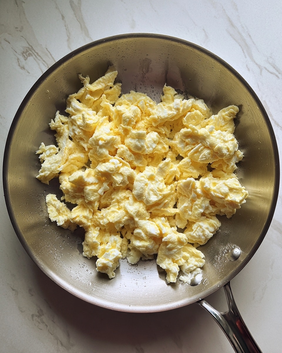 A large silver pan contains scrambled eggs with soft, fluffy clumps in pale yellow and white colors scattered loosely inside. The pan’s smooth metal surface reflects some light, showing the shiny texture and droplets of oil near the eggs. The background shows a white marbled surface with faint veins and light shadows. photo taken with an iphone --ar 4:5 --v 7