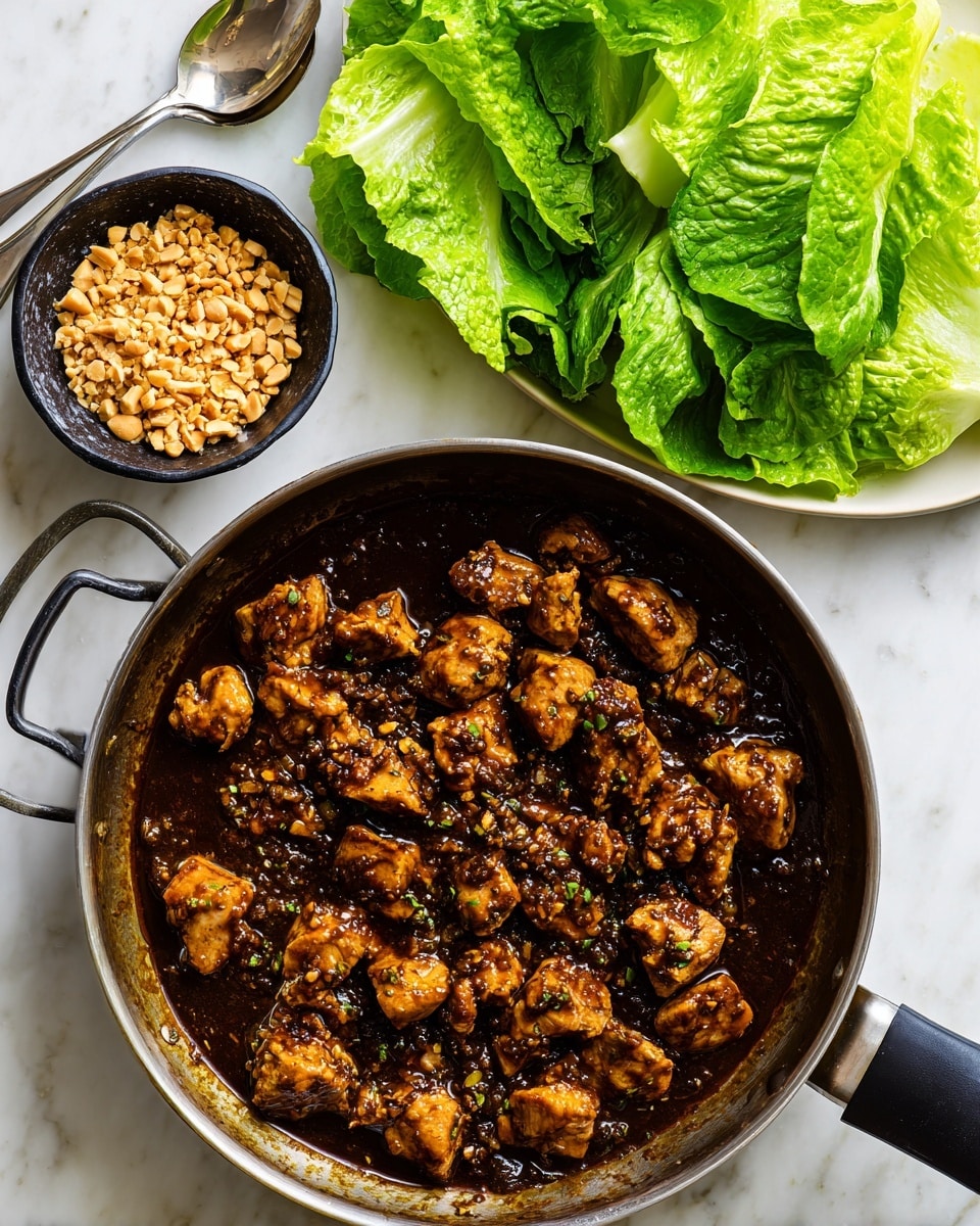 A round pan with a black handle sits on a white marbled surface, filled with small chunks of browned chicken in a thick dark brown sauce, coating and surrounding the pieces. Beside the pan is a small black bowl filled with crushed peanuts, located on the upper middle left of a rectangular white plate covered with fresh bright green butter lettuce leaves arranged loosely, some overlapping. A silver spoon rests on the top left corner of the plate. The lighting is even, highlighting the textures and colors clearly, photo taken with an iphone --ar 4:5 --v 7