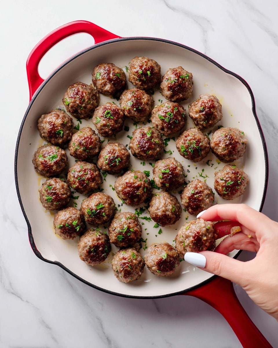 A red cast iron skillet with a white inside holds about thirty small round meatballs arranged in rows. A woman's hand with light skin and white nail polish is picking up one meatball from the skillet. The background surface is white marbled texture. photo taken with an iphone --ar 4:5 --v 7