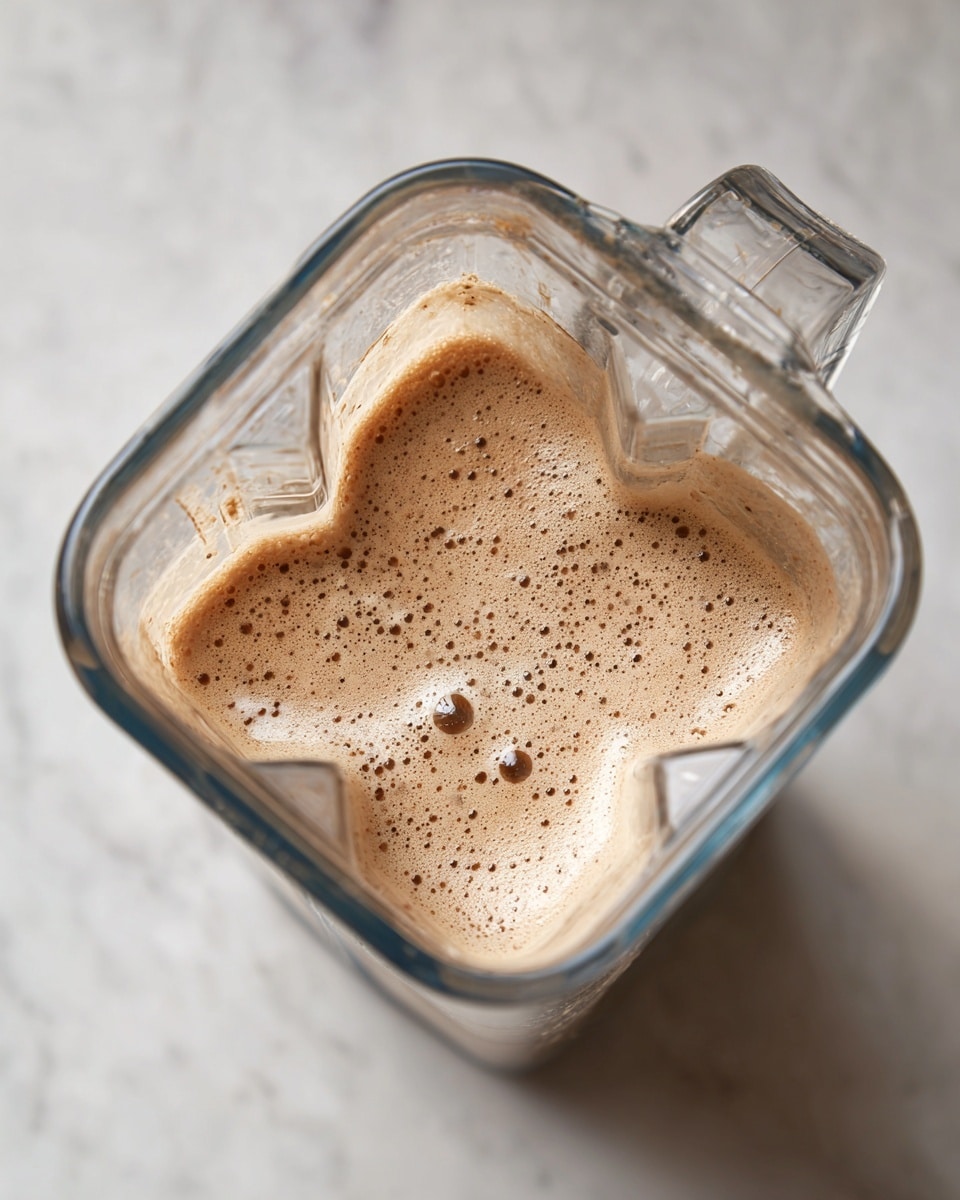 A tall clear blender container filled with a light brown blended mixture that has a slightly frothy texture on top and small dark brown specks throughout. The container is placed on a white marbled surface and is open at the top without a lid, showing the inside texture clearly photo taken with an iphone --ar 4:5 --v 7