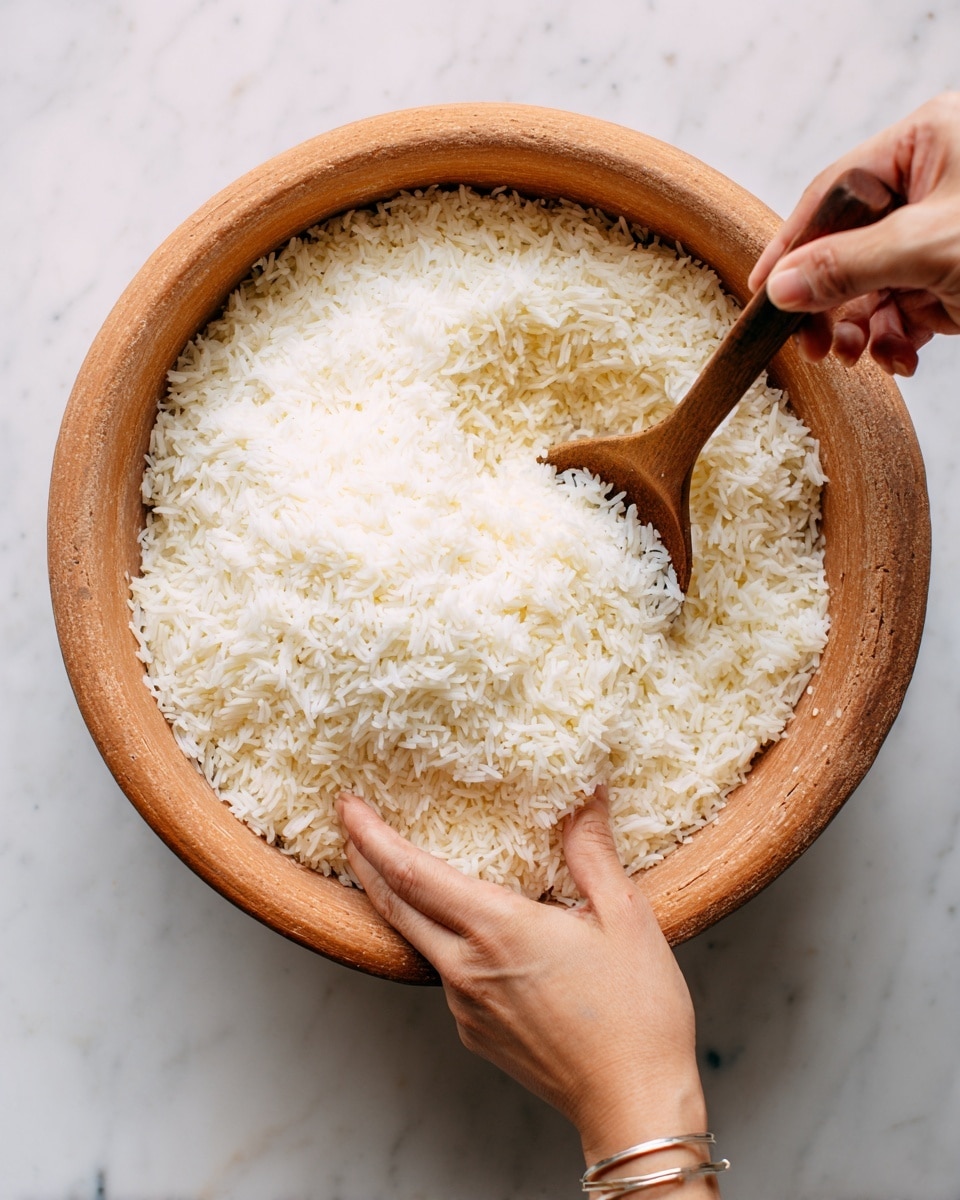 A close-up view of a round clay pot filled with a generous mound of fluffy white rice. The texture of the rice grains is soft and slightly sticky, piled high and evenly spread inside the pot. A wooden spoon rests inside the pot, angled slightly upward under the rice. A woman's hand with a simple silver bracelet on her wrist holds a small white bowl, tilting it over the rice as if adding or mixing something. The background shows a clean white marbled surface that adds a smooth, bright contrast to the warm, earthy tones of the clay pot. photo taken with an iphone --ar 4:5 --v 7