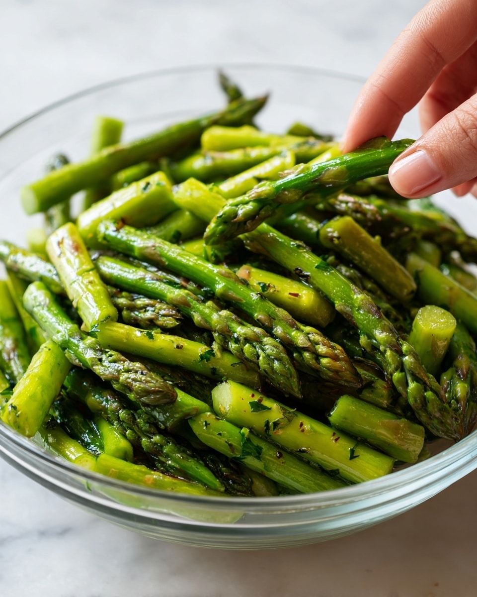 The image shows a clear glass bowl filled with bright green asparagus stalks that have been trimmed and coated in a shiny, light oil with visible herbs scattered on them. The asparagus pieces are cut into two parts, and the spears are facing different directions, giving a natural, fresh look. The bowl sits on a white marbled surface, and a woman's hand is reaching towards the top right, lightly touching some asparagus. photo taken with an iphone --ar 4:5 --v 7