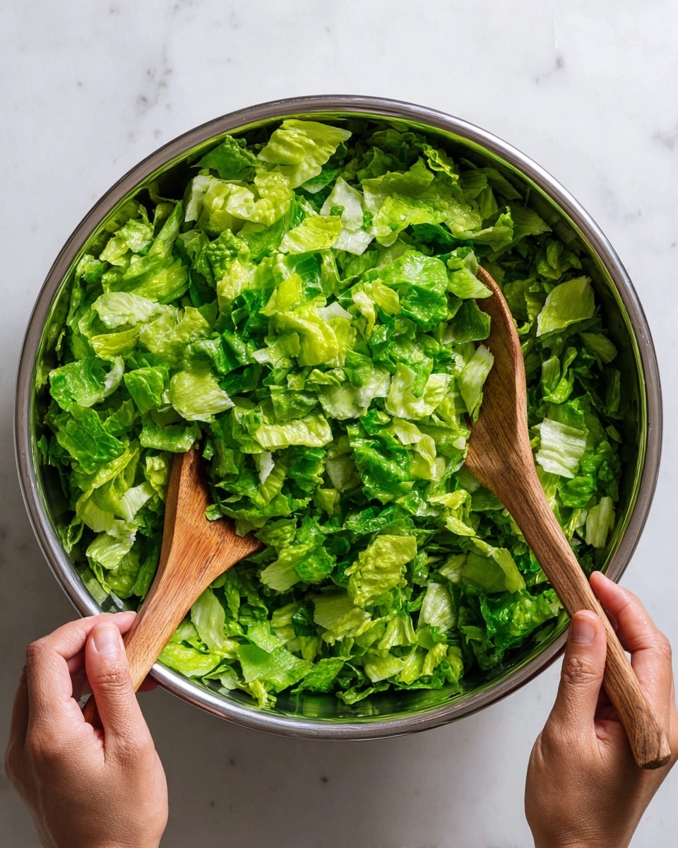 A large silver mixing bowl filled with chopped green lettuce in different shades, ranging from light green to dark green, showing fresh leafy texture. Two wooden spoons are stirring the lettuce inside the bowl, held by a woman's hand on each side, both hands visible at the edges of the image. The bowl sits on a white marbled surface, creating a clean and bright background. Photo taken with an iphone --ar 4:5 --v 7