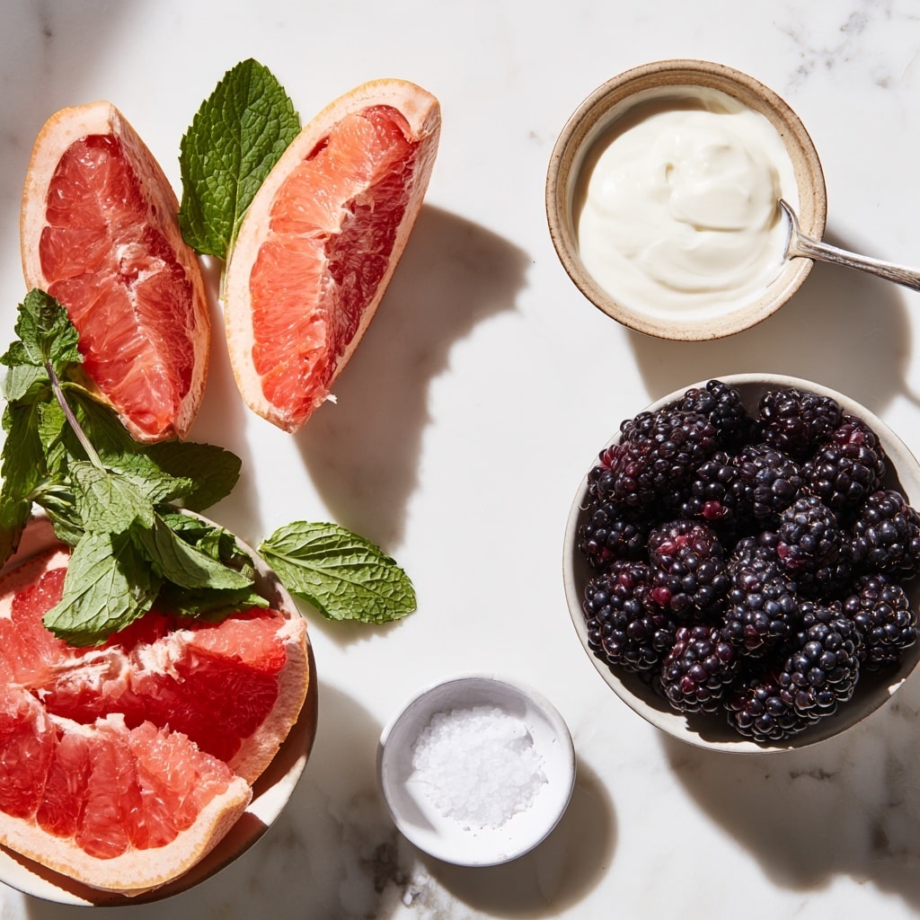 A close-up of a white bowl filled with a mix of blackberries and small pink grapefruit segments, all coated in a creamy white sauce that lightly covers the fruits and clings to their textured surfaces. The blackberries are deep purple-black and bumpy, while the grapefruit pieces show a soft pink color with a small green leaf on one piece. The bowl sits on a wooden surface with a white marbled texture background. Photo taken with an iphone --ar 4:5 --v 7