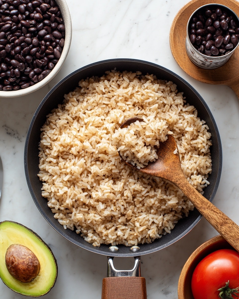 A black pan with cooked light brown rice mixed with small white onion pieces spread evenly inside, with a wooden spoon resting in the pan lifting some rice. To the left, there is a white ceramic bowl filled with black beans and a spoon inside it, and behind it, an open can of black beans. At the bottom left corner, half an avocado with a brown pit and a whole red tomato are visible. On the right side, an empty light wooden bowl sits on a white marbled surface. Photo taken with an iphone --ar 4:5 --v 7