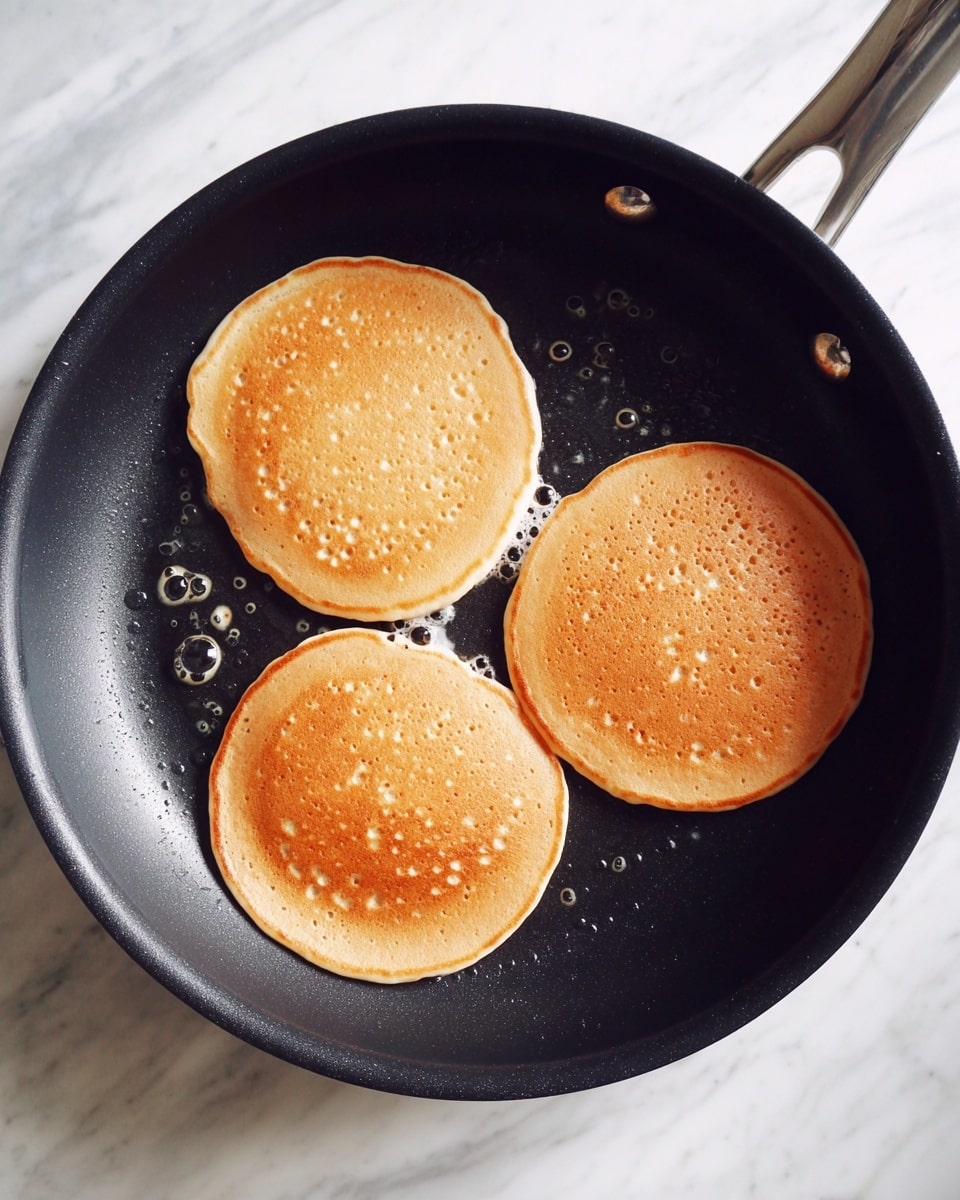 Three light tan pancake batter rounds cook on a black frying pan, showing small bubbles on their smooth, slightly shiny surfaces. The frying pan rests on a white marbled surface. photo taken with an iphone --ar 4:5 --v 7