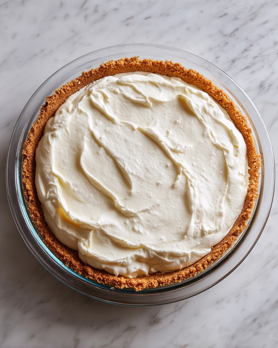A round pie with a golden brown crust forms the bottom layer, filling most of a clear glass pie dish. On top of the crust is a thick layer of smooth, white cream spread evenly all over, leaving a small border of crust visible around the edge. The pie dish sits on a white marbled surface, showing a clean and simple setting. Photo taken with an iphone --ar 4:5 --v 7