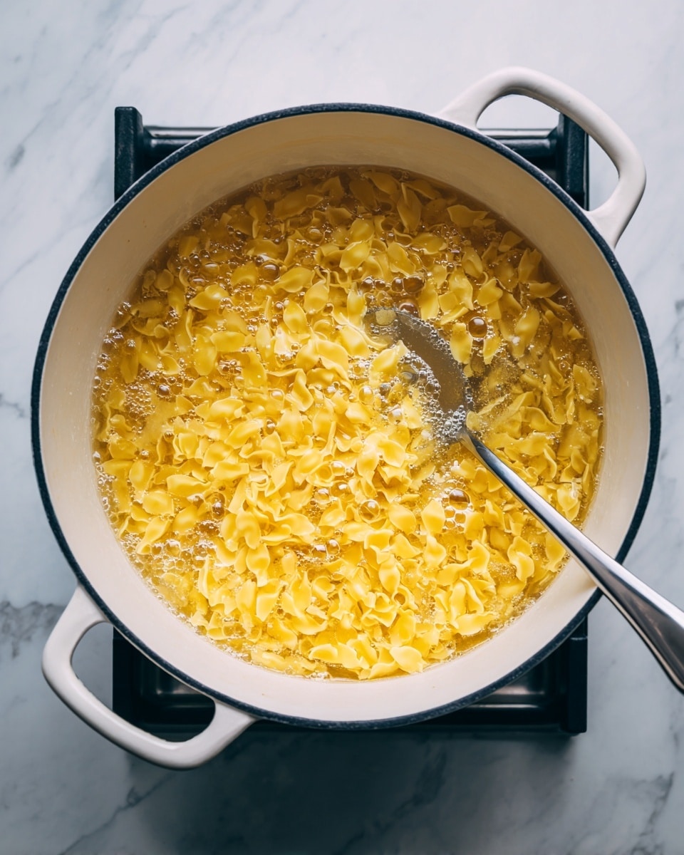 The image shows a white pot with black inside edges filled with many small, yellow pasta pieces boiling in bubbling water on a stove. A silver spoon is partially dipped into the pot, stirring the pasta on the right side. The pot handles are white, and the stove beneath has black grates. The background surface is a white marbled texture photo taken with an iphone --ar 4:5 --v 7