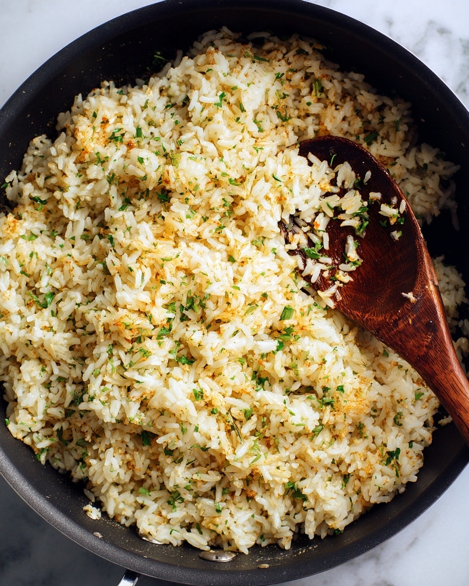 A close-up view of a black pan filled with light golden brown cooked rice mixed with small bits of green herbs scattered evenly throughout. A dark wooden spoon is stirring the rice at the right side of the pan, with the spoon's bowl partially covered by rice grains. The rice texture looks fluffy with some grains slightly separated. The background surface is a white marbled texture. photo taken with an iphone --ar 4:5 --v 7