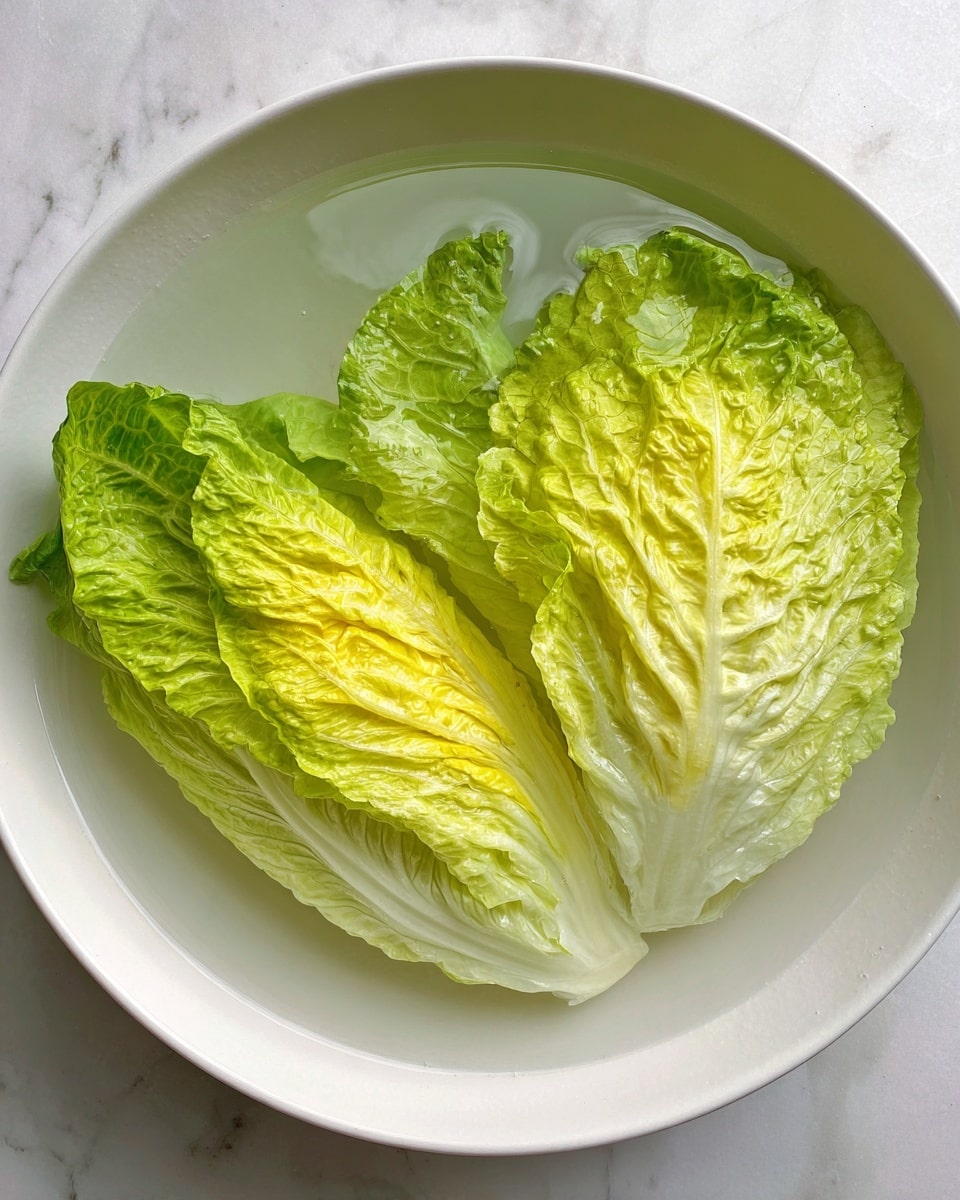 Two large leaves of pale green and bright yellow lettuce are placed flat in a white bowl filled with clear water. The leaves show detailed texture with crisp veins running through them, the outer edges lightly ruffled and a bit darker green. The bowl sits on a white marbled surface. photo taken with an iphone --ar 4:5 --v 7