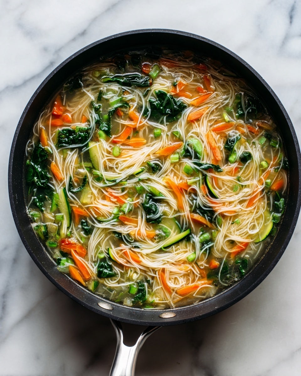 A black pan filled with a clear light broth. Floating in the broth are thin, pale white noodles, chopped orange carrots, slices of green zucchini, and bright green leafy vegetables. The vegetables and noodles are spread evenly throughout the broth, creating a colorful and fresh look. The pan is placed on a white marbled surface with its silver handle facing toward the bottom. photo taken with an iphone --ar 4:5 --v 7
