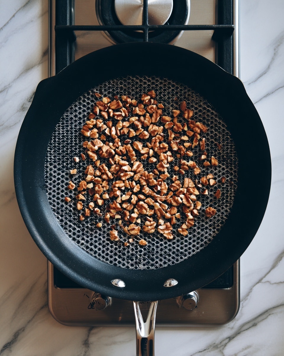 A black frying pan with a hexagon pattern on the inside holds a single layer of light brown walnut halves scattered evenly across the pan. The pan sits on a stovetop with a silver burner visible below it. The background has a white marbled texture. photo taken with an iphone --ar 4:5 --v 7