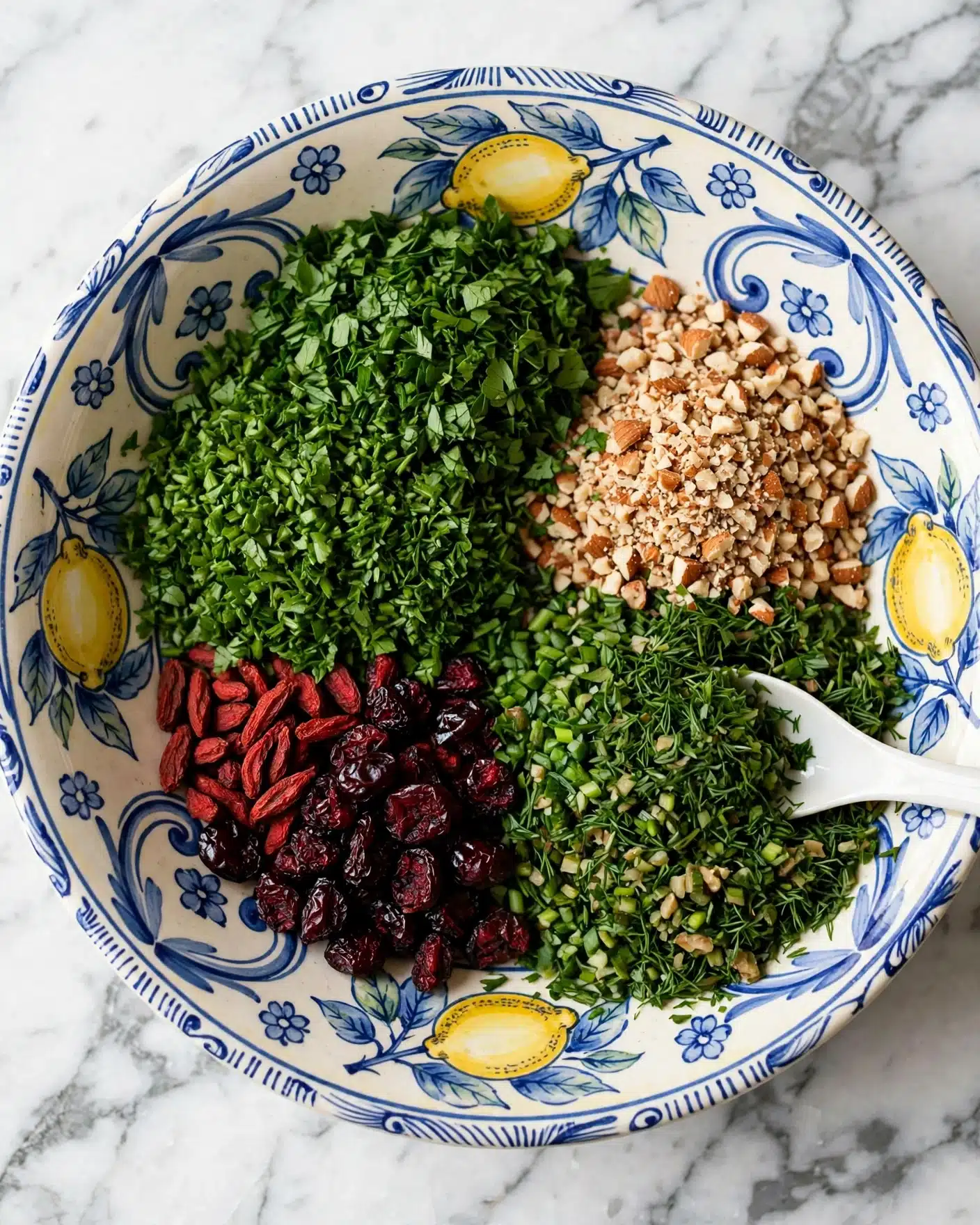 A colorful ceramic bowl with a lemon and blue pattern holds four main layers of ingredients arranged separately: a large section of finely chopped bright green herbs, a pile of small chopped nuts in light brown, a cluster of dark red dried berries, and a pile of more mixed green chopped herbs. A white spoon is partly buried in the green herbs on the right side. The bowl rests on a white marbled surface. photo taken with an iphone --ar 4:5 --v 7