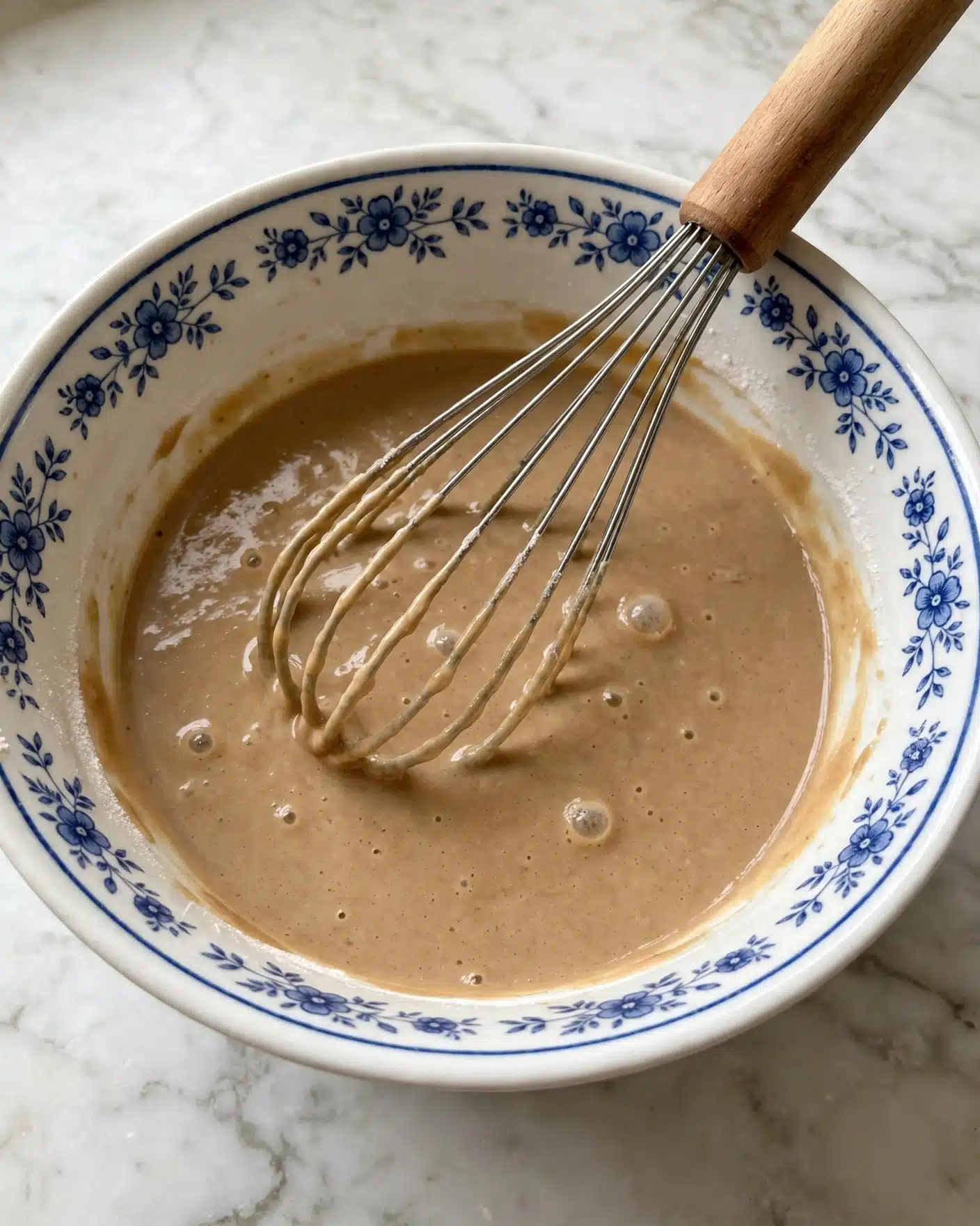 A white bowl with a blue flower pattern on the rim holds a light brown, smooth batter with a few bubbles on the surface. A metal whisk with a wooden handle rests diagonally inside the bowl, partially coated with the batter. The bowl is placed on a white marbled surface. photo taken with an iphone --ar 4:5 --v 7