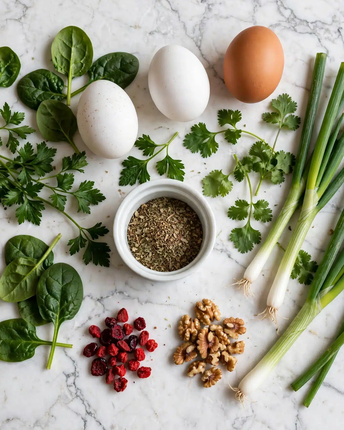 The image shows a white marbled surface with fresh ingredients spread out neatly: three whole eggs, two white and one brown, surrounded by green leafy herbs including spinach, parsley, cilantro, and green onions. Near the center is a small white dish filled with dried herbs. Below the eggs are small piles of red dried berries and chopped walnuts, adding texture and color contrast to the green herbs and white eggs. photo taken with an iphone --ar 4:5 --v 7