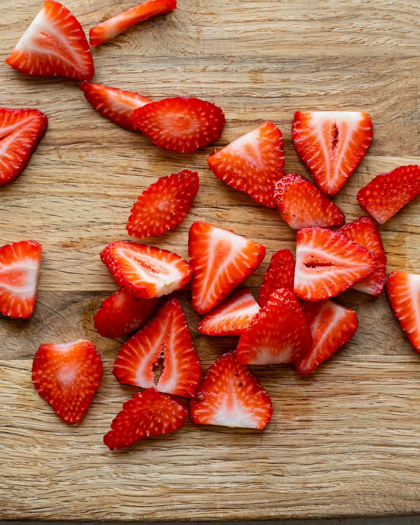 Strawberry Tiramisu (Creamy, Light and Fresh) Recipe 4 The image shows many strawberry slices spread out on a wooden cutting board. The slices vary in size and shape, with most being triangular or oval. The strawberries are bright red with visible seeds and some white inner parts near the top edges. The wooden cutting board has a light brown color with visible grain texture. photo taken with an iphone --ar 4:5 --v 7