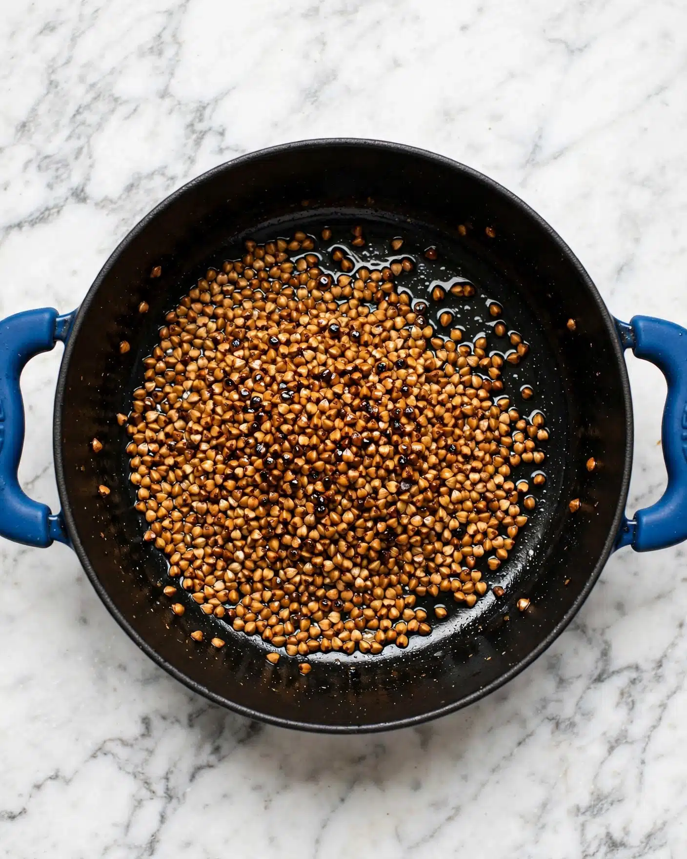 A top-down view of a black pot with blue handles on both sides, placed on a white marbled surface. Inside the pot, a single visible layer of small grain-like pieces that are toasted to a golden brown with some darker spots, scattered unevenly but mostly gathered towards the center. The grains have a shiny texture, likely from a little oil. The number 