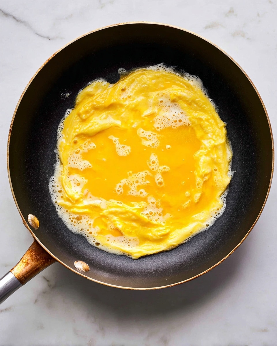 The image shows a top view of a black frying pan with a shiny golden handle, containing a single, thin layer of bright yellow-orange scrambled eggs cooking inside. The eggs have a slightly wet texture with small bubbles forming on the surface and some white and light yellow patches where the eggs are more cooked. The edges of the eggs are slightly crispy and lacy, sticking a little to the pan's sides. The pan is placed on a white marbled surface. photo taken with an iphone --ar 4:5 --v 7