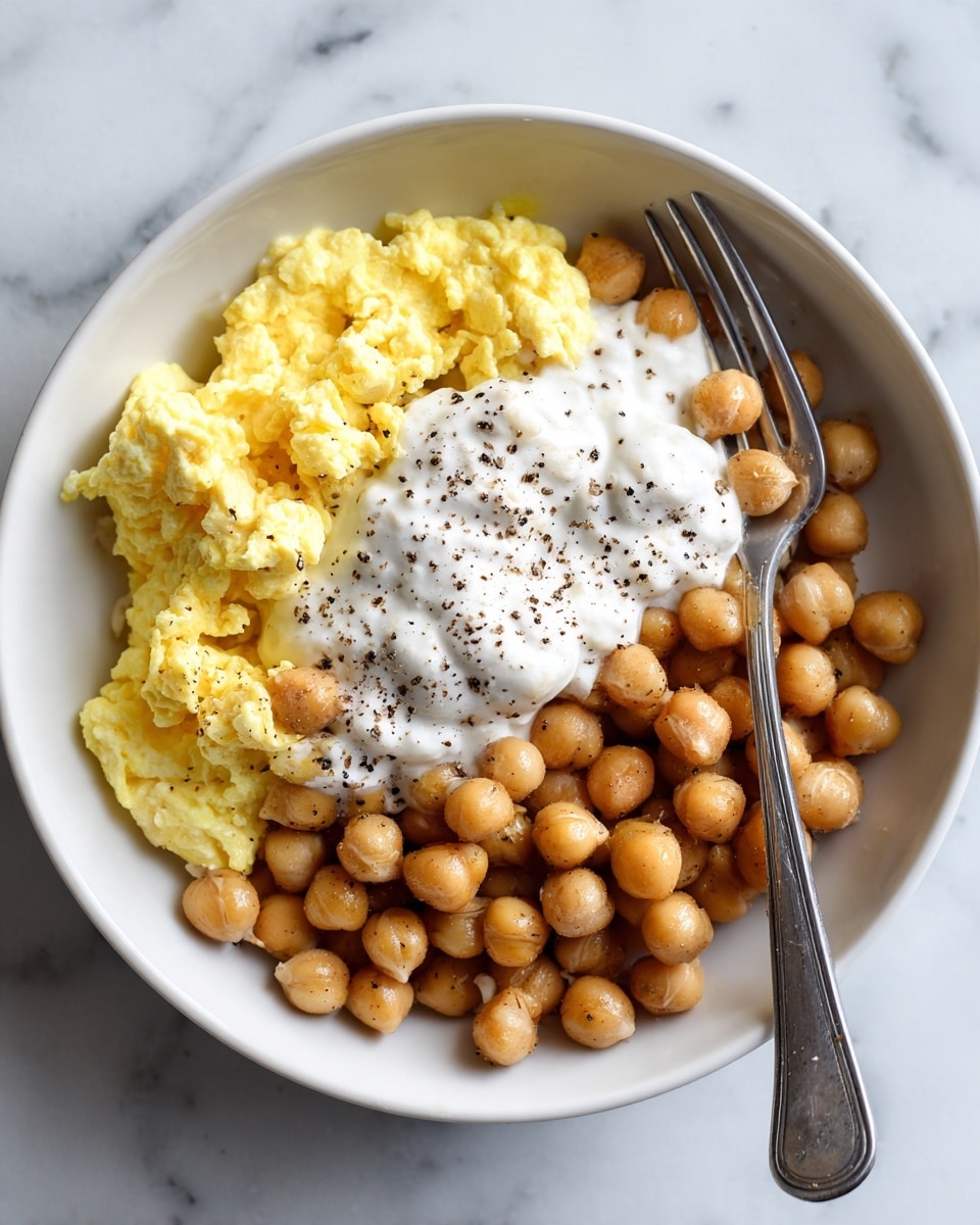 The image shows a white bowl with three main layers inside: scrambled eggs that are yellow and fluffy fill one side, creamy white yogurt or sauce is spread in the center with some black pepper sprinkled on top, and plump, light brown chickpeas cover the other side. A silver fork rests diagonally over the eggs and chickpeas. The bowl is placed on a white marbled surface. photo taken with an iphone --ar 4:5 --v 7