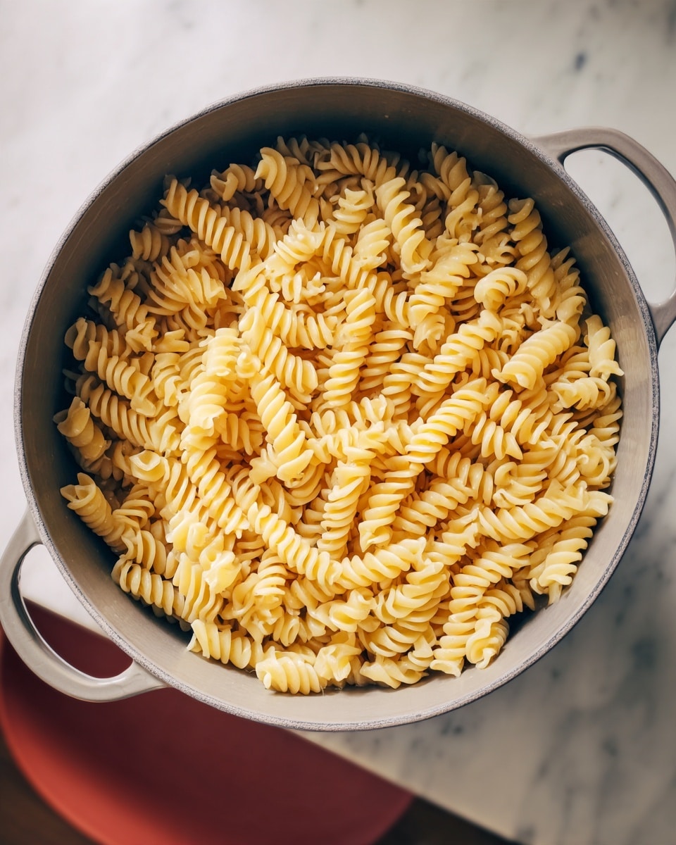 A large gray cooking pot filled with one layer of light yellow spiral pasta with a firm texture, resting on a white marbled surface. The pasta pieces are tightly packed, showing their ridged spiral shape clearly. The pot has small side handles, and part of a reddish chair is visible in the background. Photo taken with an iphone --ar 4:5 --v 7