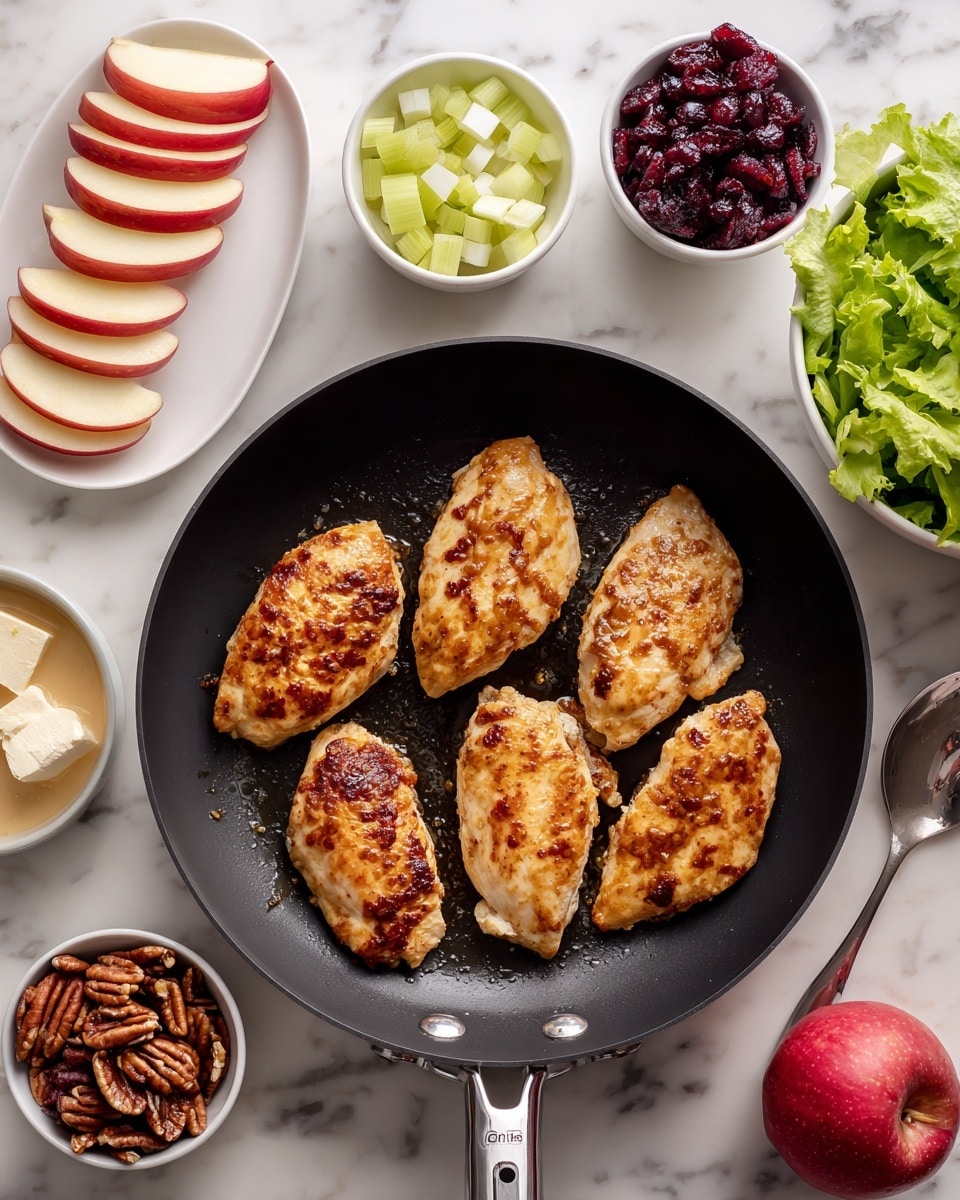 A black frying pan with five golden-brown cooked chicken pieces spread out evenly inside it, showing a slightly crispy texture with some darker brown spots. Around the pan, there are small white bowls arranged on a white marbled surface, each with different ingredients: to the top left, thin red apple slices fanned out on a white plate; near the top center, diced pale green celery; next to it, thin slices of light purple onion; to the right, dark red dried cranberries; below the cranberries, crumbly white cheese; at the bottom left, glazed pecans in a white bowl; beside the pecans, a bowl with light beige dressing and a spoon; and at the bottom right, a bowl of fresh mixed green lettuce leaves. A whole red apple is placed near the bottom right side of the image. Photo taken with an iphone --ar 4:5 --v 7