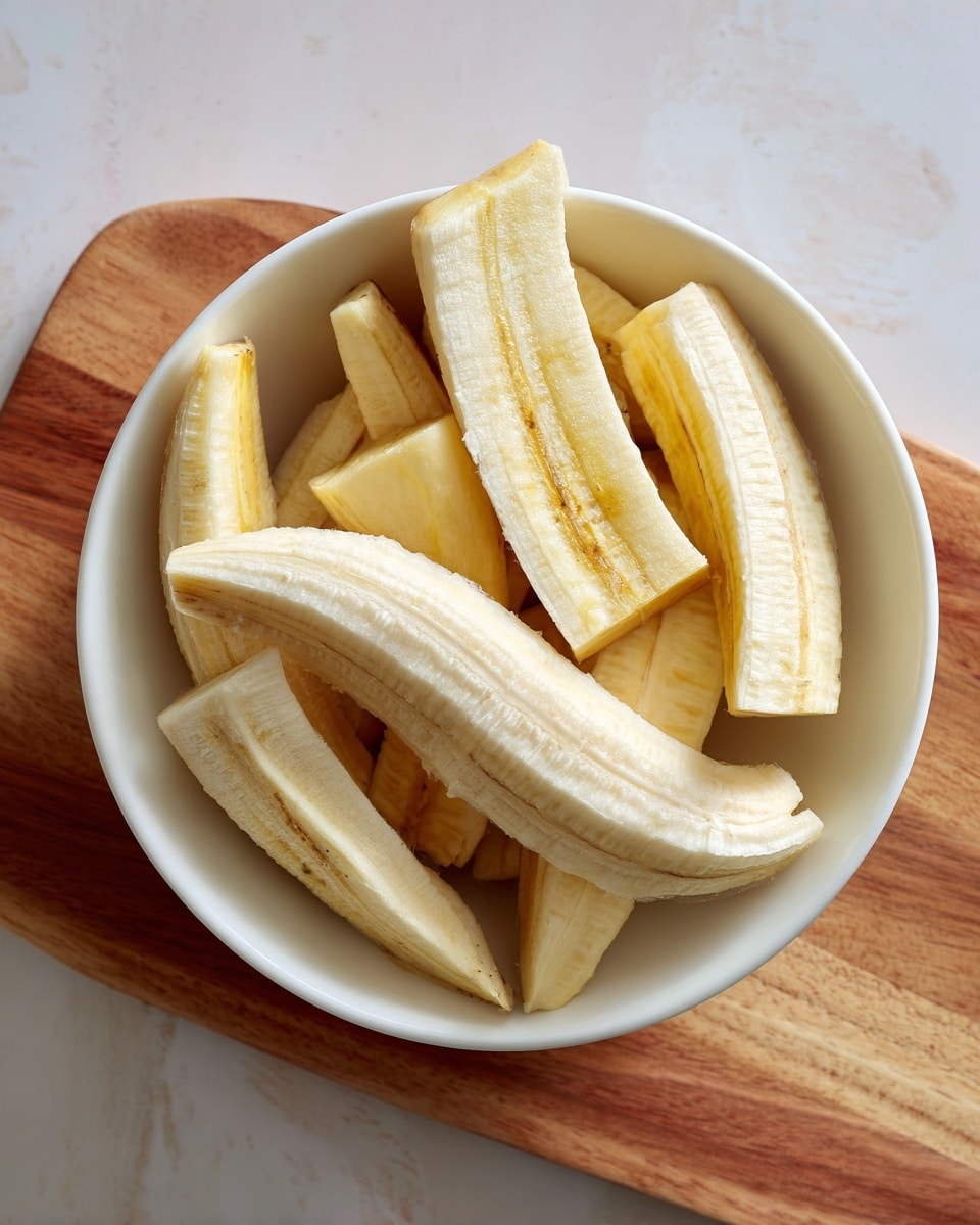 A white textured bowl filled with several pieces of peeled yellow plantains cut into large chunks, with some pieces showing a smooth, slightly fibrous texture and others having visible ends and curving edges, all resting on a wooden board against a white marbled texture background photo taken with an iphone --ar 4:5 --v 7