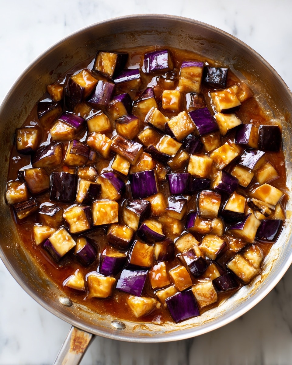 A metal pan filled with small, uneven cubes of cooked eggplant showing dark purple skin and soft, light yellow flesh. The eggplant pieces are covered in a glossy, thick brown sauce that pools slightly at the bottom of the pan, giving a shiny and moist look. The pan sits on a white marbled surface with a power cord visible on the side. photo taken with an iphone --ar 4:5 --v 7