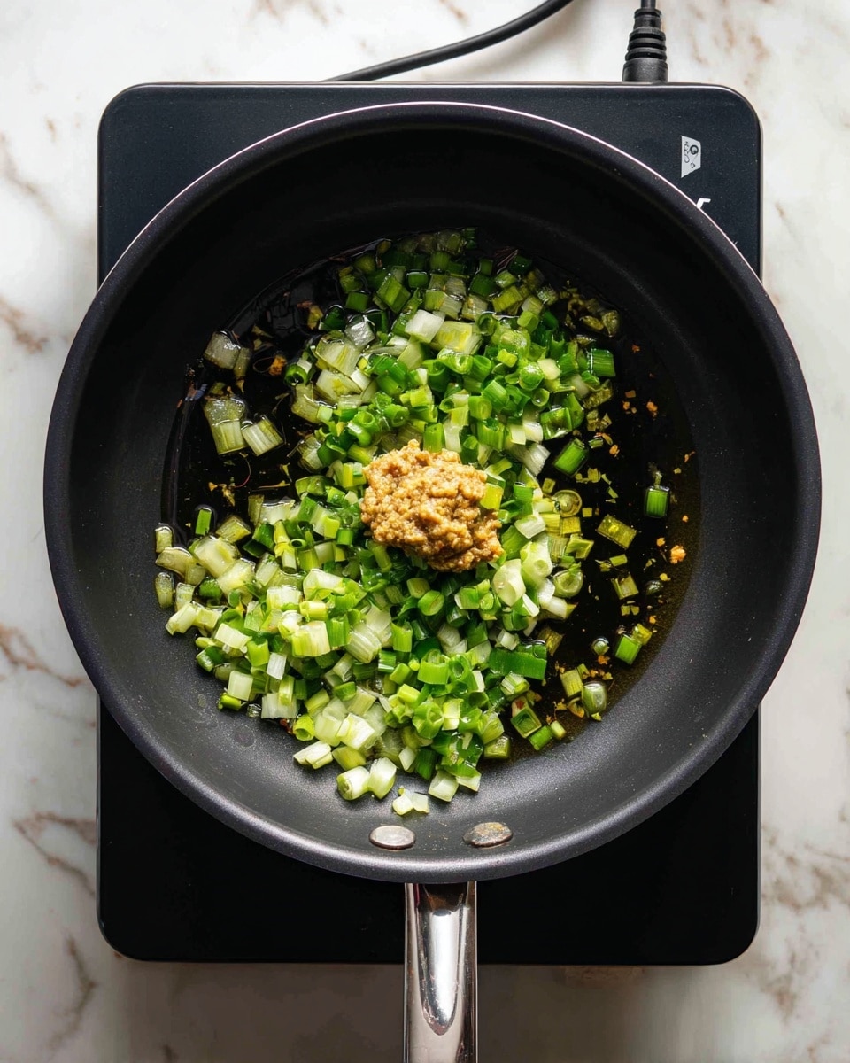 Inside a black cooking pot, there are small pieces of light green chopped vegetables scattered around the base with some oil glistening on the surface. In the center, there is a dollop of light brown paste sitting on top of the vegetables. The pot is placed on a black stove with a white cord visible in the background, and the whole scene is set on a white marbled texture. photo taken with an iphone --ar 4:5 --v 7
