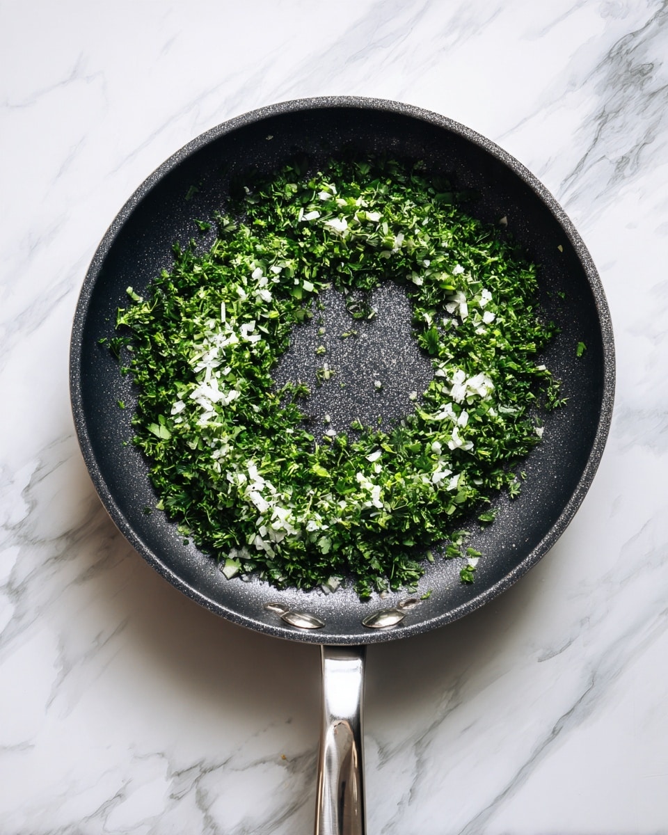 A dark frying pan with a silver handle sits on a white marbled surface. Inside the pan, there is a single layer of finely chopped bright green herbs and small white pieces, spread evenly in a circular shape but leaving parts of the pan's black surface visible. The herbs and white pieces have a fresh, moist texture. The photo is taken with an iphone --ar 4:5 --v 7