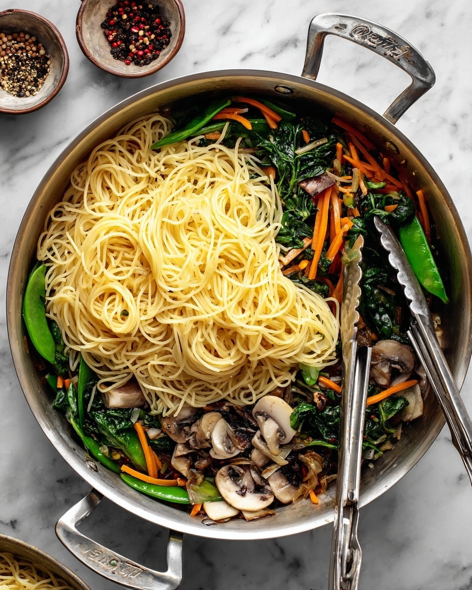A shiny silver pan filled with three layers: the bottom layer has mixed cooked vegetables including green snow peas, dark green spinach leaves, white and brown mushroom slices, and thin strips of orange carrot, showing a mix of smooth and slightly crunchy textures. On top of that, there is a thick pile of light yellow cooked spaghetti noodles, soft and long, covering most of the vegetables but some are visible around the edges. Two silver tongs rest inside the pan, one on the right side and the other on the bottom, placed diagonally over the noodles. The pan sits on a white marbled surface with small bowls of black pepper and chili flakes placed next to it on the top left. Photo taken with an iphone --ar 4:5 --v 7