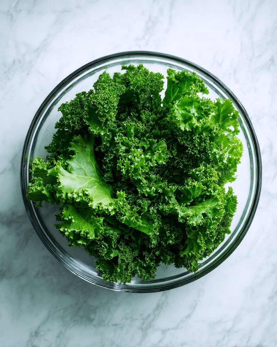 A clear glass bowl is filled with fresh, bright green kale leaves. The kale has a mix of smooth and curly textures, with some light reflection showing its freshness. The bowl sits on a white marbled surface that adds a clean and bright backdrop to the rich green color of the kale. photo taken with an iphone --ar 4:5 --v 7