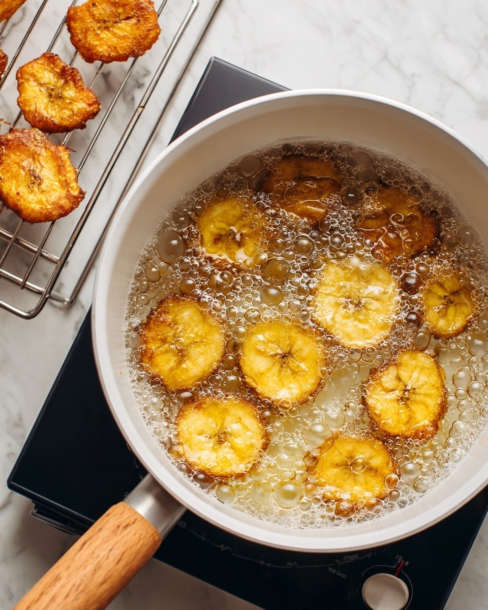 A white pot filled with golden-yellow round plantain slices frying in bubbling hot oil. The oil is full of small and large bubbles covering the surface. The pot has a light wooden handle and sits on a black electric cooker with a visible dial. To the left, on a metal cooling rack placed over a pan, some fried plantain slices sit, showing a crispy texture with rough edges and a mix of golden and light brown spots. The background is a white marbled surface. photo taken with an iphone --ar 4:5 --v 7