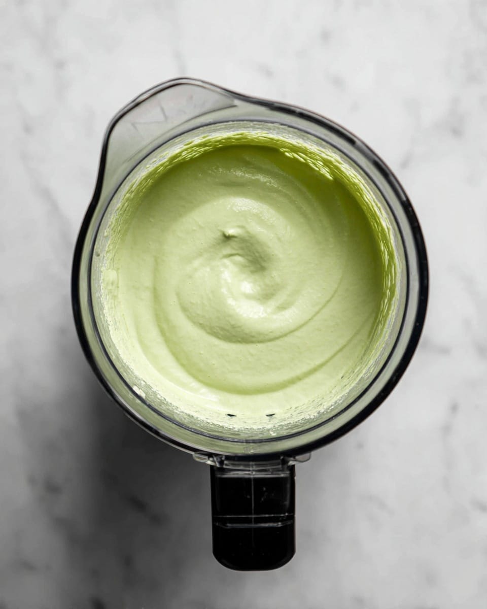 The image shows a clear blender container placed on a white marbled surface, filled with a smooth, light green creamy mixture. The mixture is thick and evenly blended, filling about half of the blender's depth. The blender container has a black lid attachment on one side, and the container's edges and bottom are slightly visible through the translucent material. The surface around the blender is clean with subtle grey veining typical of white marble. Photo taken with an iphone --ar 4:5 --v 7