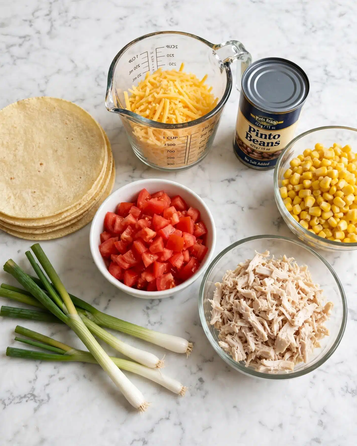 The image shows a collection of ingredients arranged neatly on a white marbled surface. There are four soft corn tortillas stacked on the left side. In front of the tortillas, there are fresh green onions with long white bulbs and green tops. A white bowl filled with bright red diced tomatoes is placed near the center. To the right of the tomatoes, a clear glass bowl holds shredded cooked chicken in pale beige chunks. Behind these, there is a clear measuring cup filled with shredded yellow cheddar cheese. Next to the cheese is a can labeled 