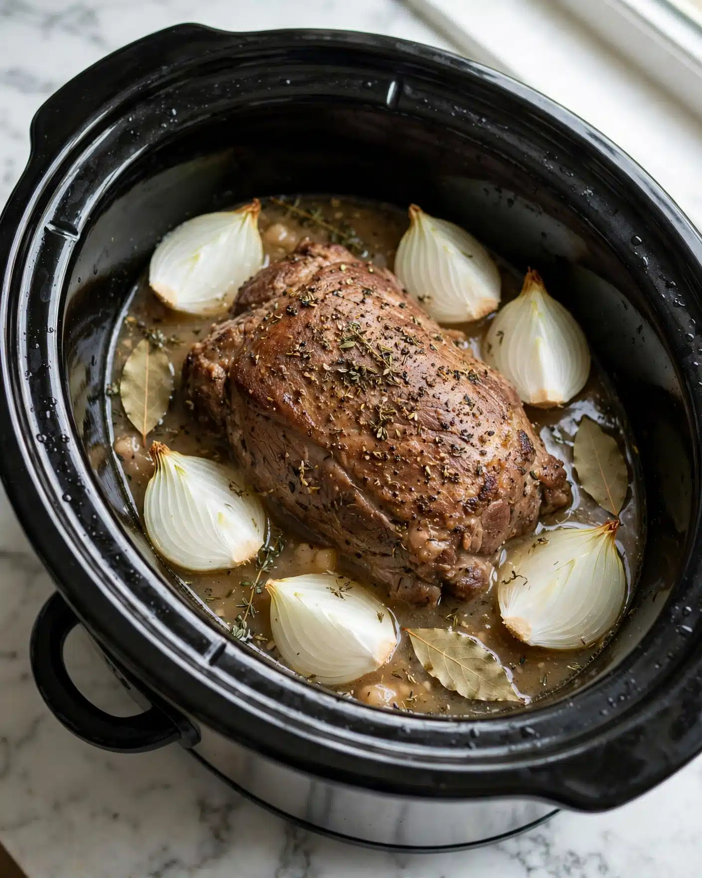 A slow cooker contains one large cooked piece of meat with a browned texture and visible seasoning, surrounded by several quartered white onion pieces that have a smooth, layered look. The meat sits in light brown liquid with some herbs sprinkled on top, and a few bay leaves rest on the surface. The slow cooker itself is black and shiny, placed on a white marbled surface. photo taken with an iphone --ar 4:5 --v 7