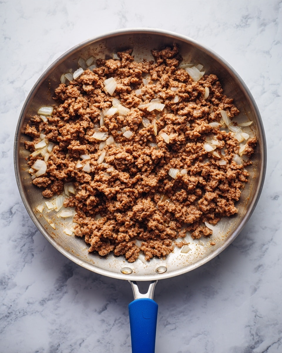 A top-down view of a silver frying pan with a blue handle, placed on a white marbled surface. Inside the pan, there is a single layer of cooked ground meat mixed with small pieces of cooked white onion. The meat is brown and crumbly, with the white onions scattered evenly throughout. The pan itself is round with a shallow depth, and the blue handle extends directly downward in the center of the image. Photo taken with an iphone --ar 4:5 --v 7