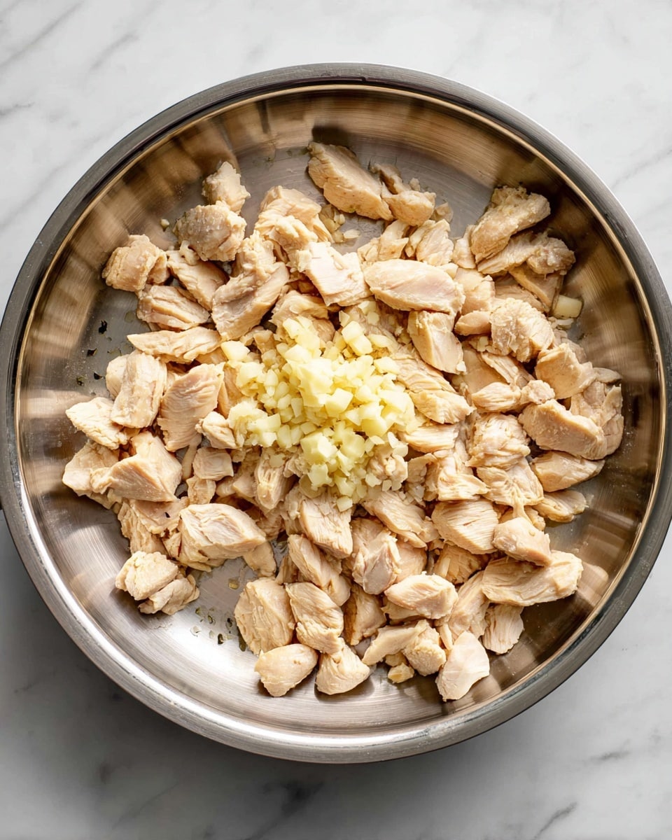A round stainless steel pan filled with small, light brown pieces of cooked chicken spreading across the pan in one layer. In the center-right of the pan, there are two small piles of finely chopped pale yellow garlic sitting on top of the chicken pieces. The pan rests on a white marbled surface. photo taken with an iphone --ar 4:5 --v 7