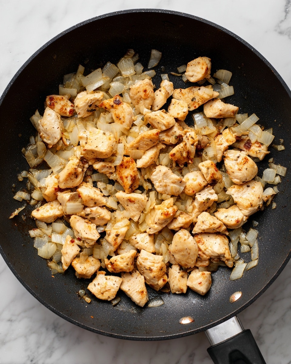 In a black frying pan on a white marbled surface, there are many small pieces of light golden chicken scattered roughly evenly across the pan. Mixed with the chicken are translucent diced onions that are slightly browned on the edges, adding a mix of white and light brown colors. The chicken pieces show a soft texture with a light sear on one side, while the frying pan has a smooth, matte black inside. The overall scene captures the cooking process mid-way, with the ingredients spread out in a casual but clear way. photo taken with an iphone --ar 4:5 --v 7