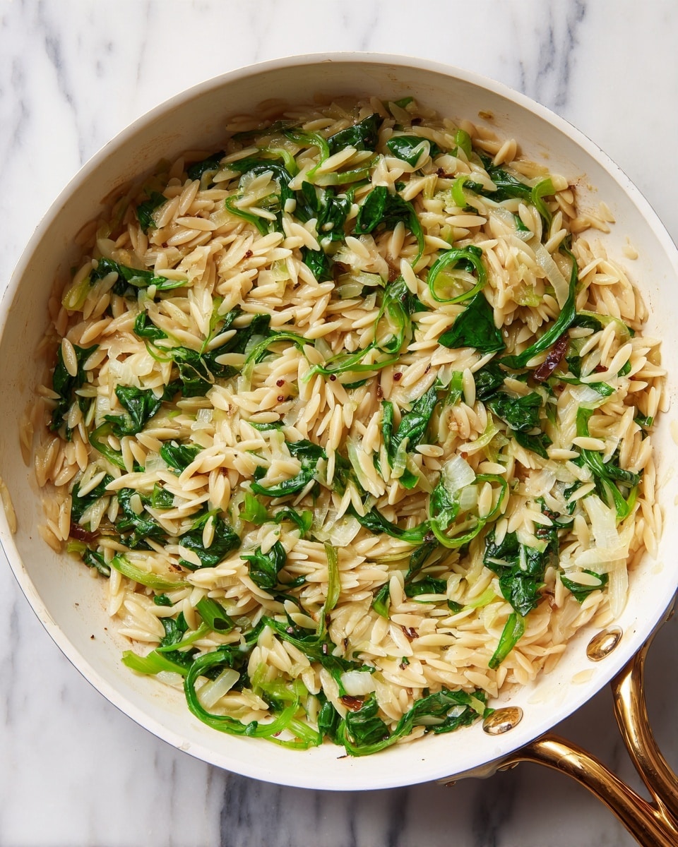 The image shows a white pan filled with a creamy dish made of light brown orzo pasta mixed with wilted dark green leafy vegetables and translucent cooked onions. The pasta grains appear soft and coated in a light sauce, while the green leaves are scattered evenly throughout. The onions are thinly sliced and spread throughout the orzo. The pan has a gold handle and is placed on a white marbled surface. Photo taken with an iphone --ar 4:5 --v 7