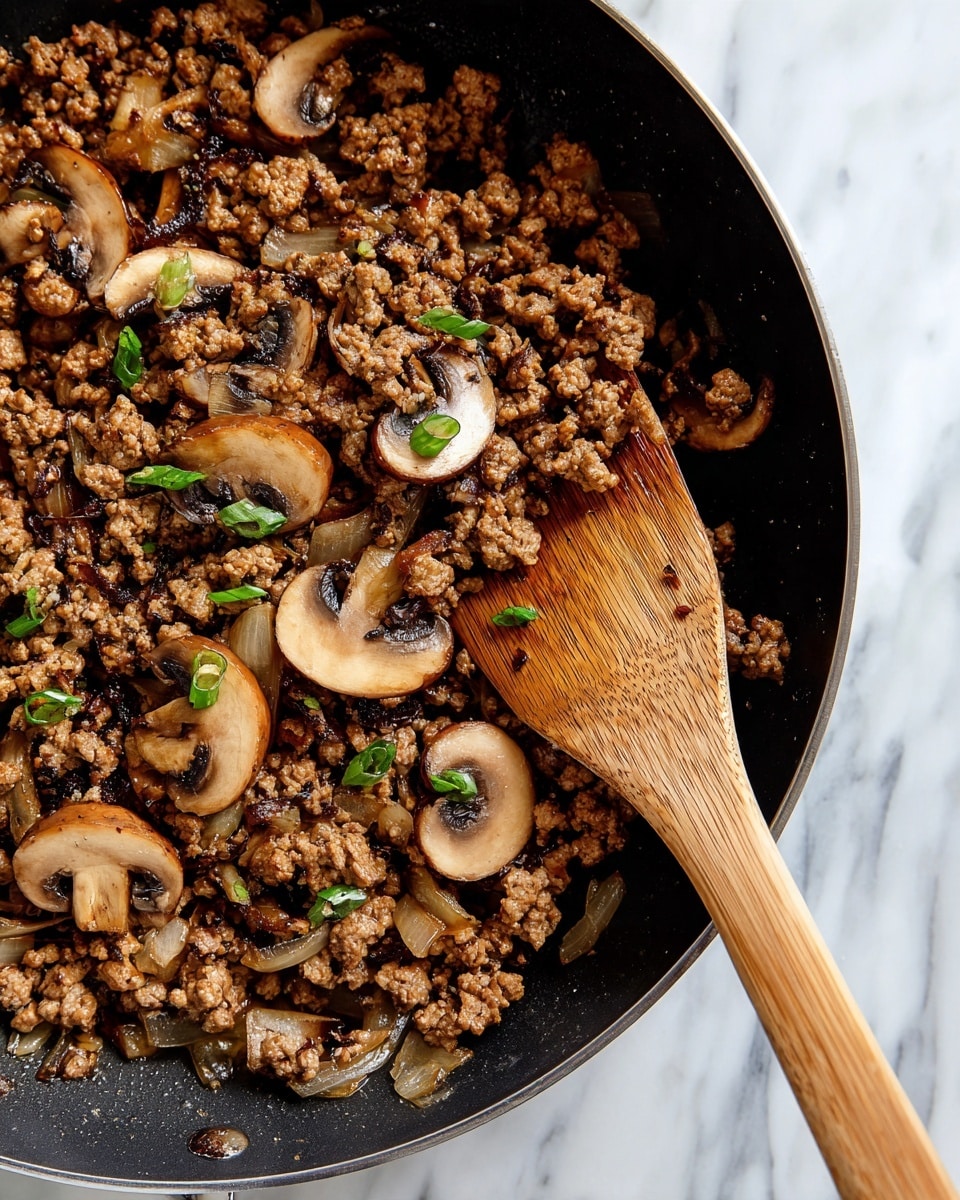 A close-up view of a black pan filled with cooked ground meat mixed with sliced light brown mushrooms and small bits of translucent cooked onions. The ingredients are well combined and lightly browned, with a few small green herb pieces scattered on top. A wooden spatula with a natural wood grain pattern is resting in the pan on the right side, slightly mixing the contents. The pan sits on a white marbled surface, adding contrast to the dark pan and food photo taken with an iphone --ar 4:5 --v 7