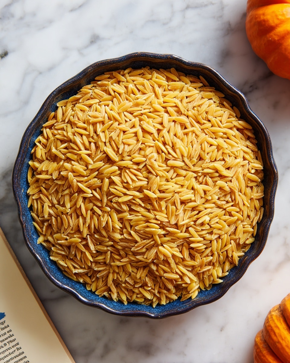 A close-up top view of golden brown toasted orzo pasta filling a dark scalloped bowl placed on a white marbled surface. The orzo is evenly spread over the entire bowl, showing a dry texture with no visible sauce or other ingredients mixed in. The edges of the bowl frame the pasta tightly, and the surrounding area includes a small orange pumpkin and part of a white and blue magazine or newspaper. Photo taken with an iphone --ar 4:5 --v 7